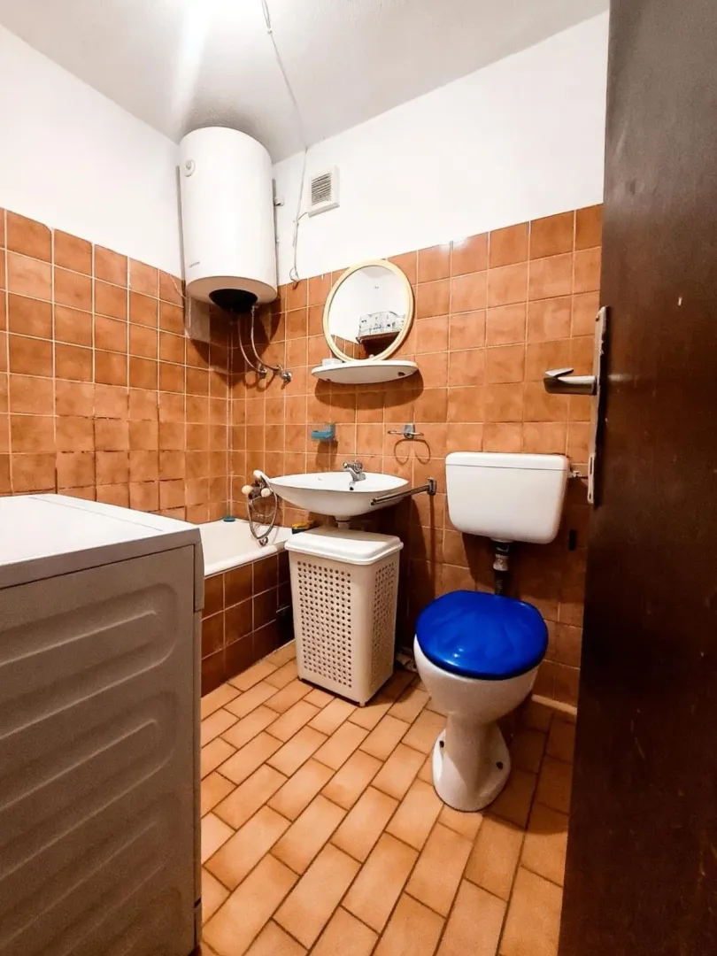 Manje kupatilo sa kadom, umivaonikom i wc šoljom obloženo braon pločicama.