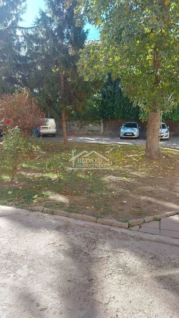 Zelena površina ispred zgrade sa drvećem i parkingom.