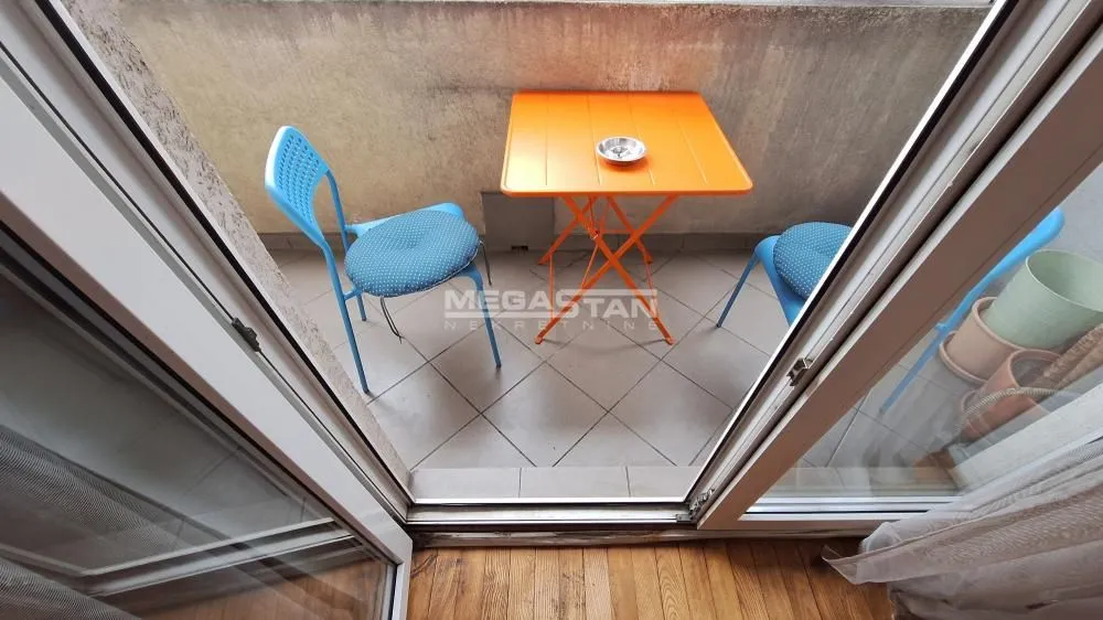 Manji balkon sa narandžastim stolom i dve stolice, gledan kroz balkonska vrata.