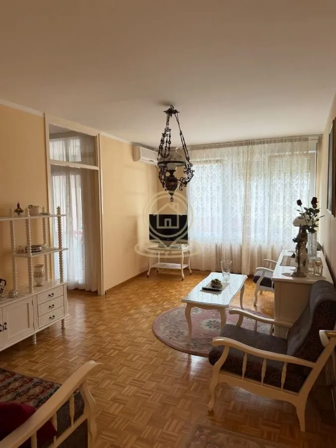 Dnevna soba sa garniturom za sedenje, parketom na podu i velikim prozorom.
