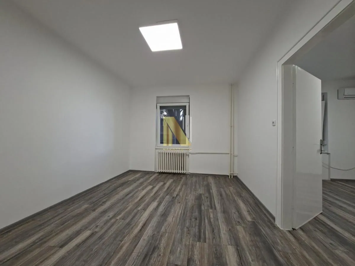Prazna soba sa belim zidovima, tamnim laminatom i jednim prozorom.