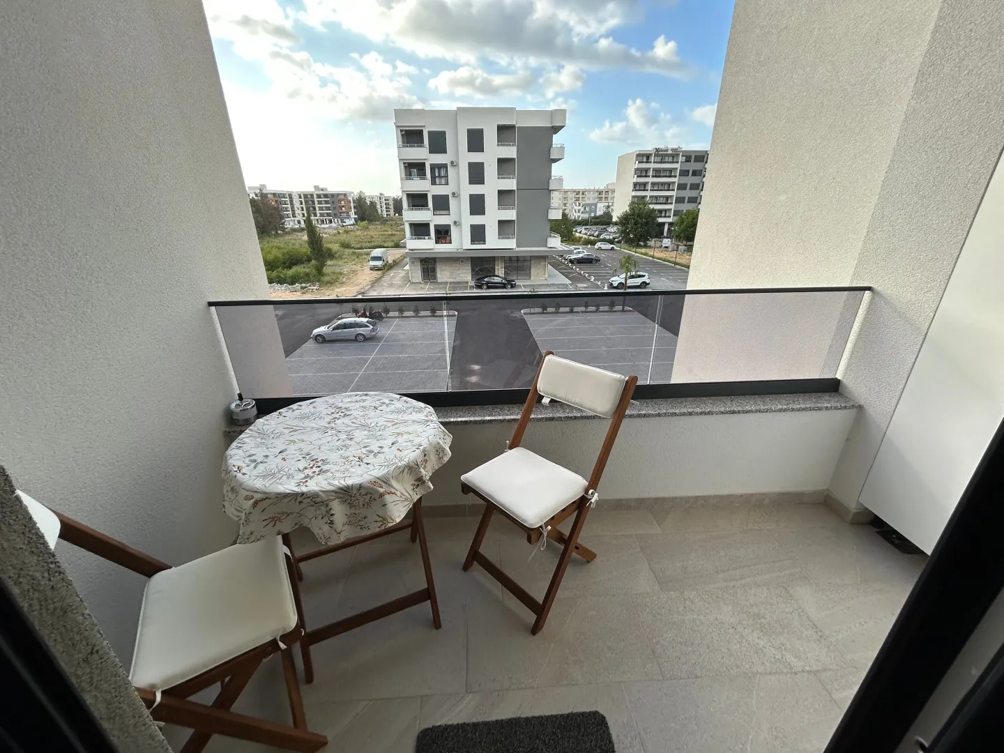 Manji namješteni balkon sa stolom i dvije stolice i pogledom na parking i zgrade.