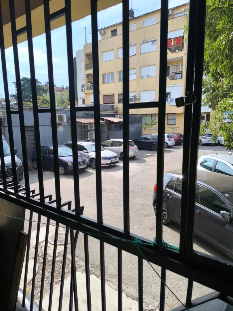 Pogled kroz zaštitne rešetke na dvorište sa parkingom i okolne zgrade.