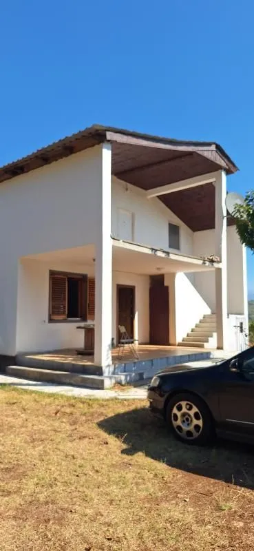 Pogled na spoljašnjost kuće sa tremom i parkiranim automobilom u dvorištu.
