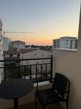 Small balcony with a black table and chair, overlooking surrounding buildings and sunset. - image 7
