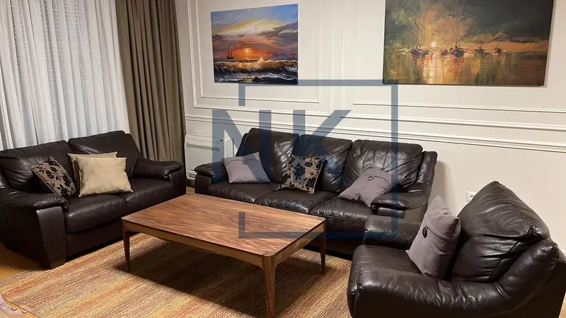 Dnevna soba opremljena tamnim kožnim garniturama, drvenim stočićem i slikama na zidu.