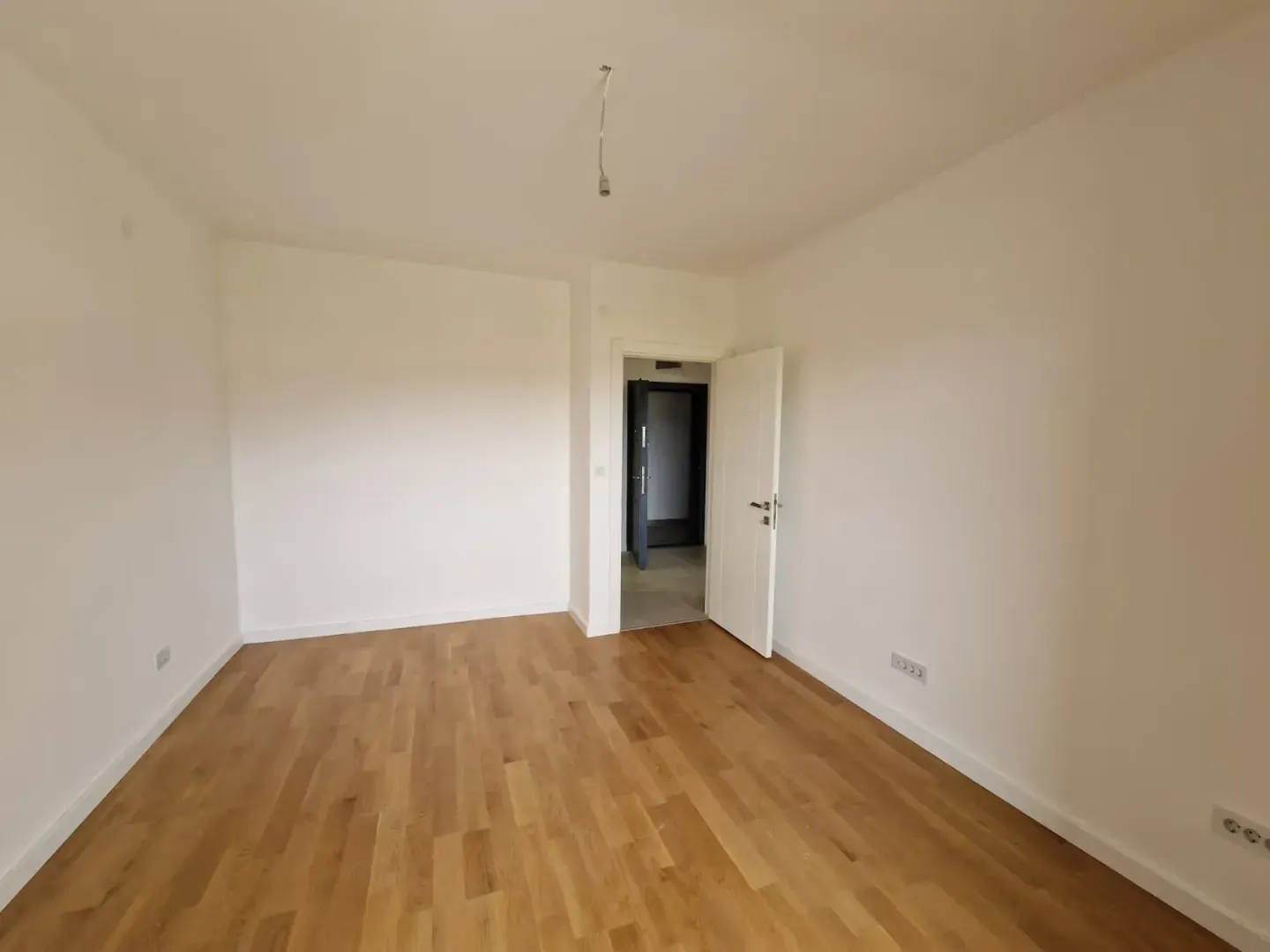 Prazna soba sa bijelim zidovima, drvenim podom i otvorenim vratima.