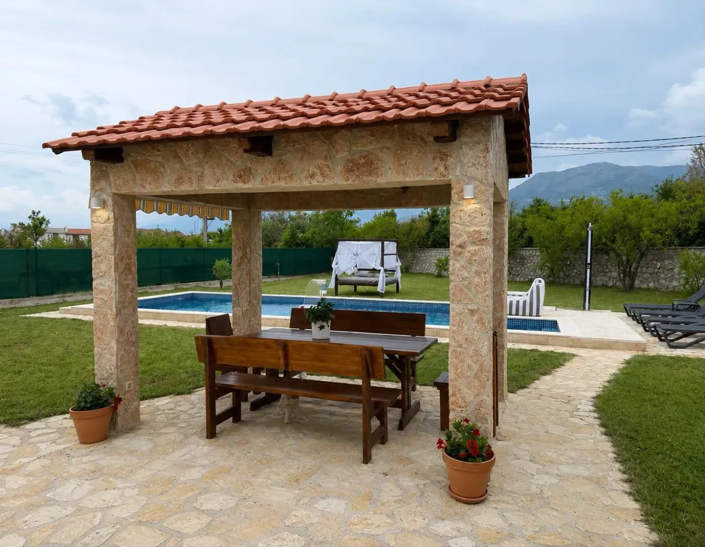 Covered patio with a wooden table and benches in a yard with a swimming pool.