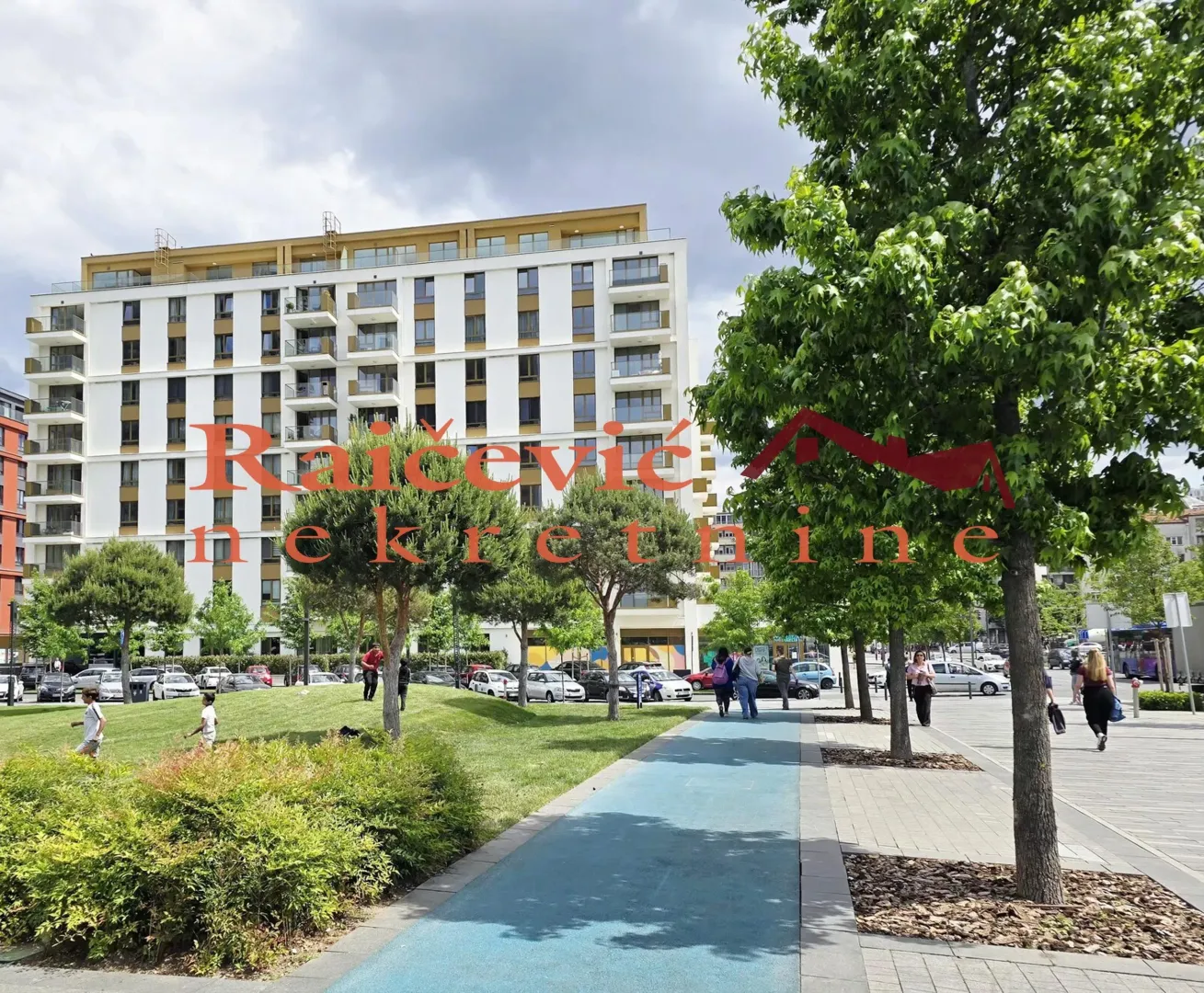 View of a modern multi-story building with landscaped green space and a walking path in the surroundings.