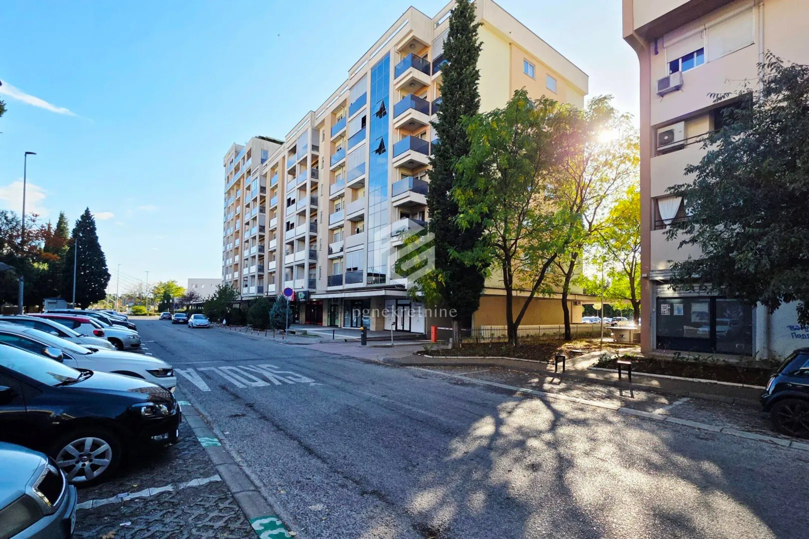 Pogled na višestambenu zgradu sa ulicom i parkiranim automobilima.