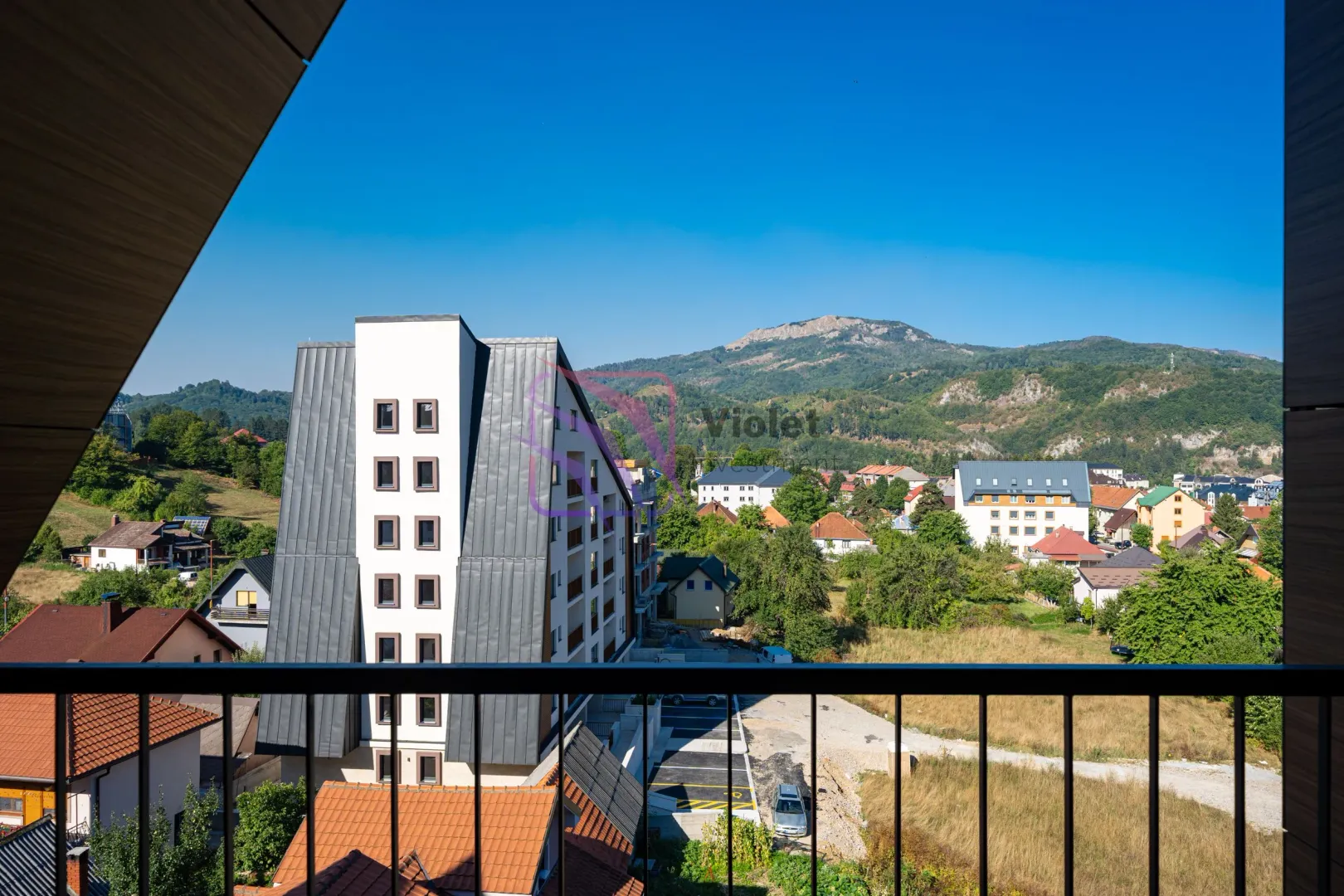 Pogled sa terase na okolne zgrade, zelene površine i planine u pozadini.