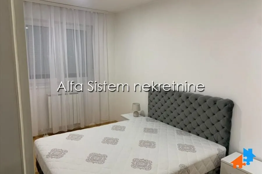 Nameštena spavaća soba sa bračnim krevetom i prozorom prekrivenim zavesama.