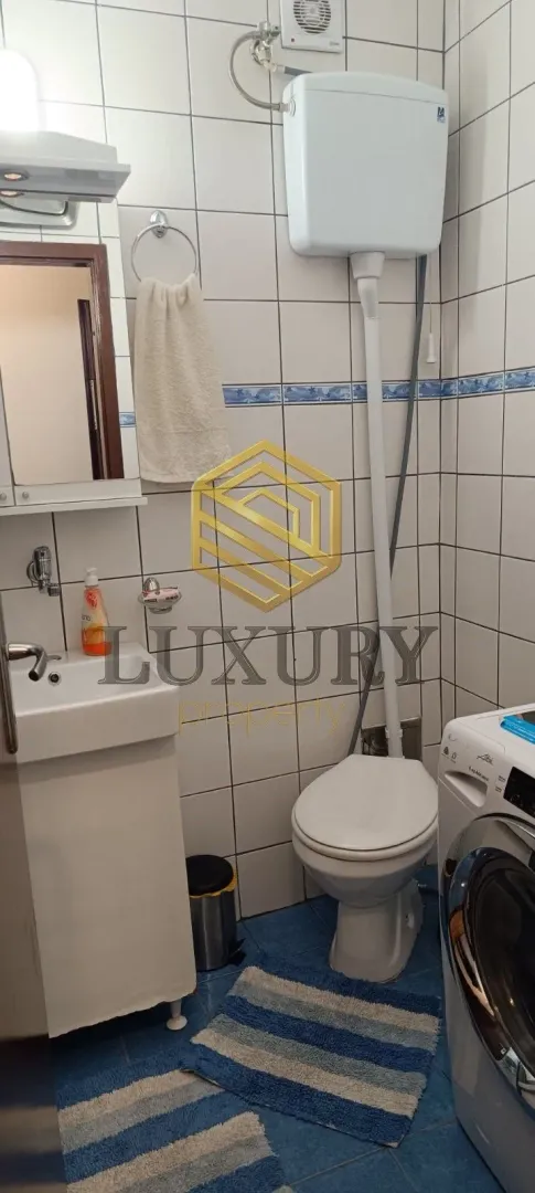 Manje kupatilo sa bijelim pločicama, umivaonikom, wc šoljom i veš mašinom.