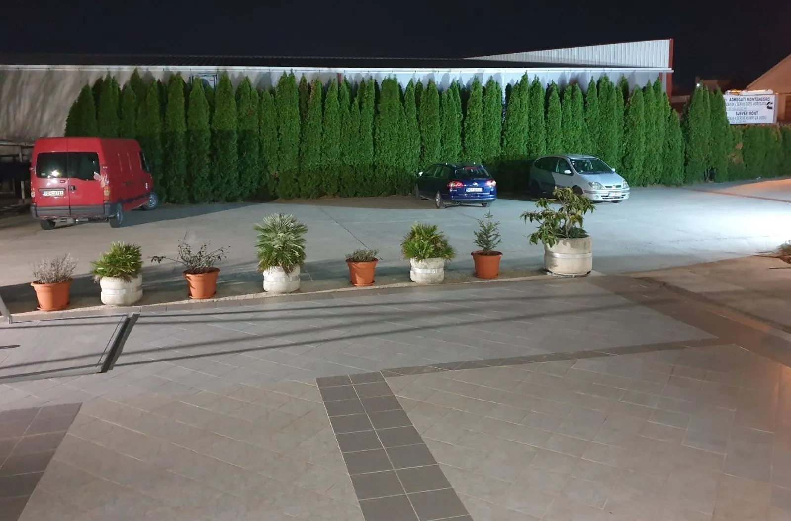 Illuminated paved yard with parking and potted plants at night.
