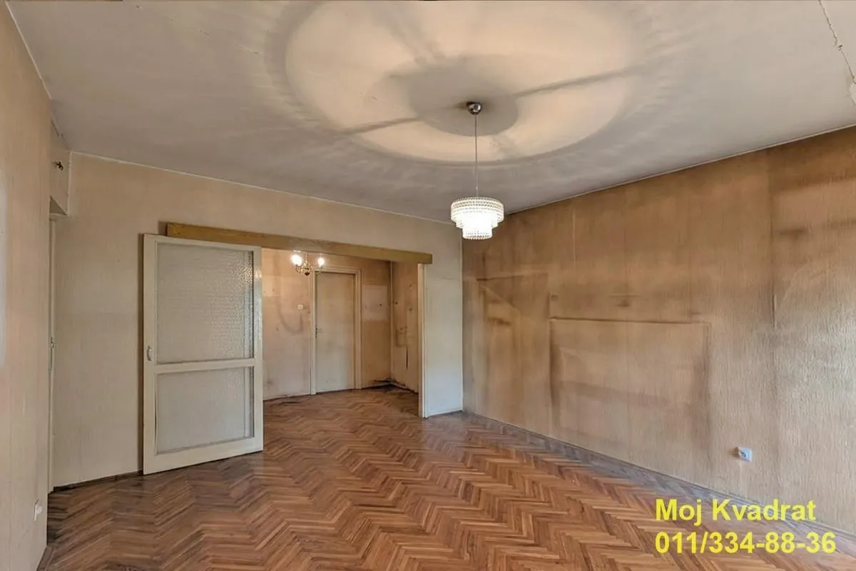 Prazna soba sa drvenim parketom i otvorenim prolazom ka drugoj prostoriji.