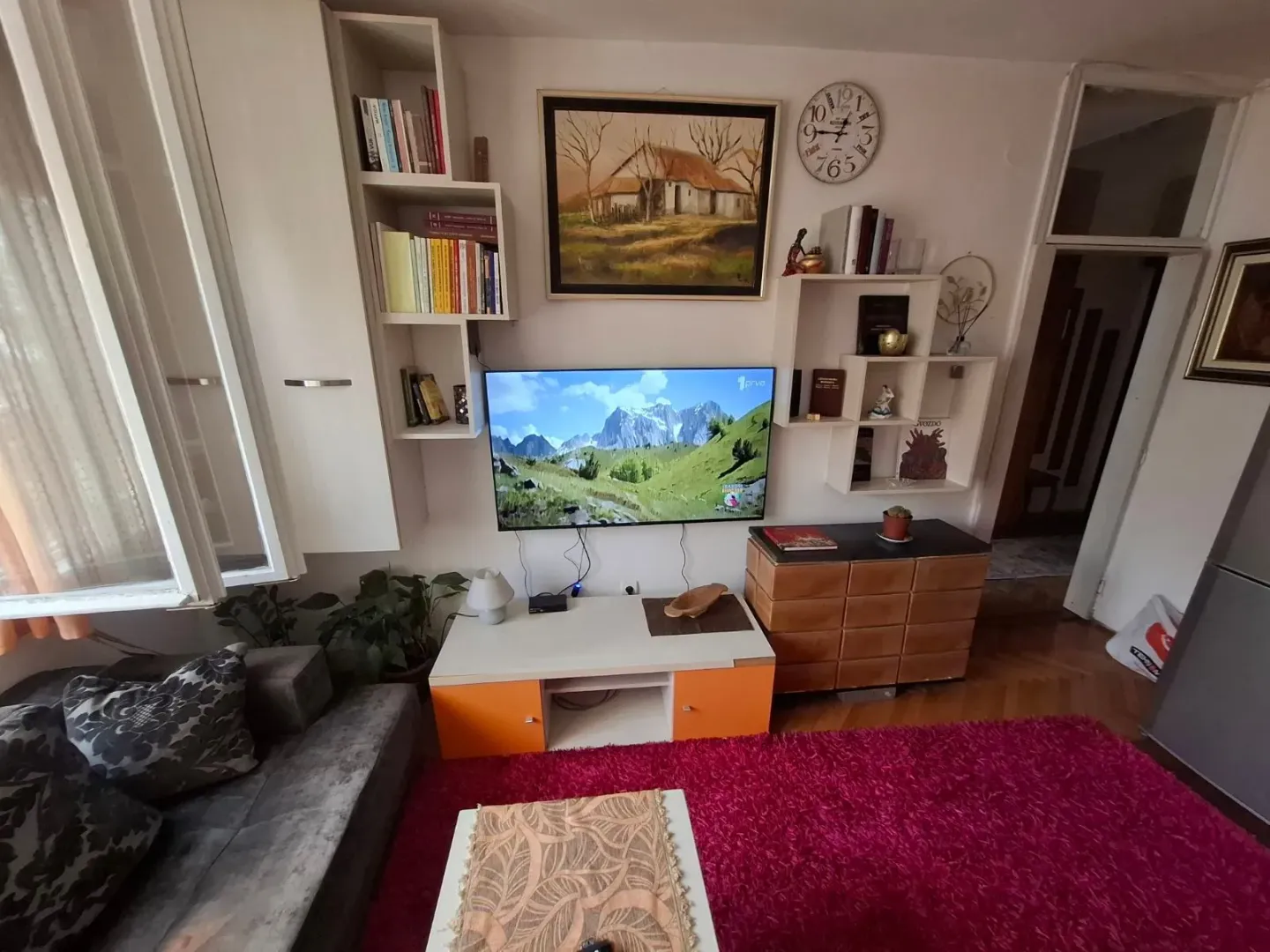 Dnevna soba sa garniturom, TV-om i zidnim policama.
