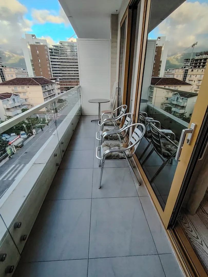 Uski popločani balkon sa staklenom ogradom, garniturom za sjedenje i pogledom na grad.