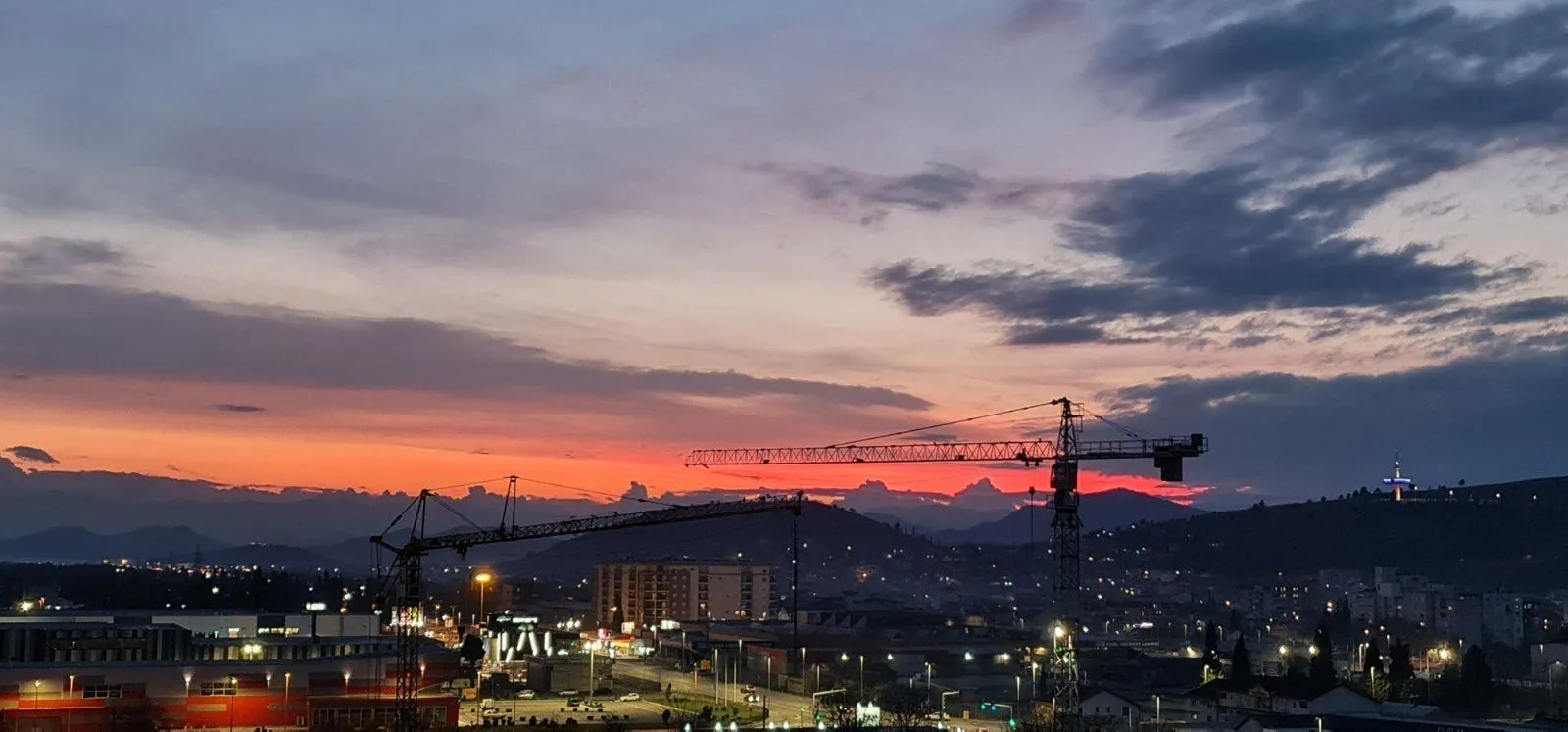 Pogled na grad i zalazak sunca sa građevinskim kranovima u daljini.