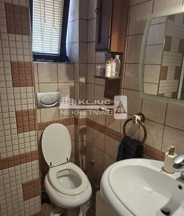 Manje kupatilo sa keramičkim pločicama, toaletom i umivaonikom sa ogledalom.