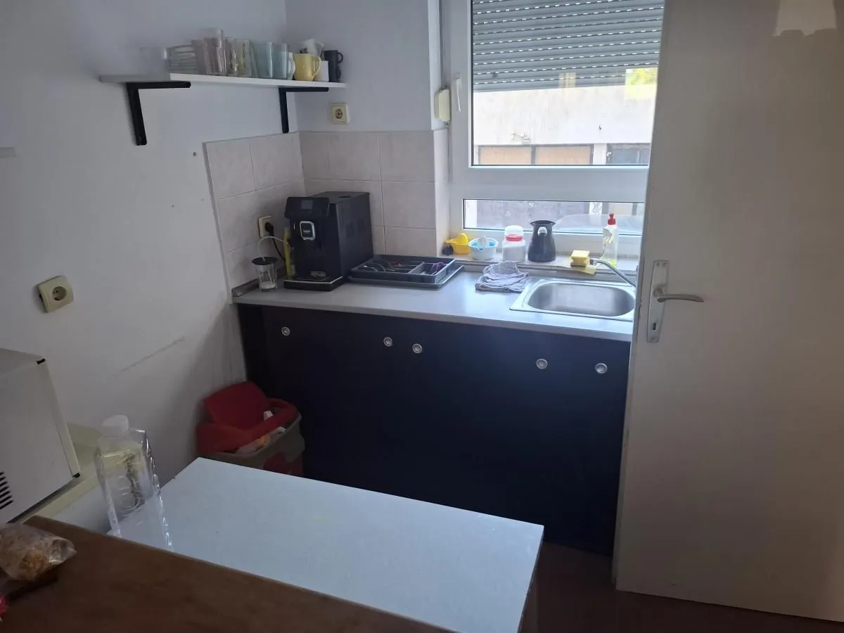 Manja kuhinja sa sudoperom, radnom površinom i aparatom za kafu pored prozora.