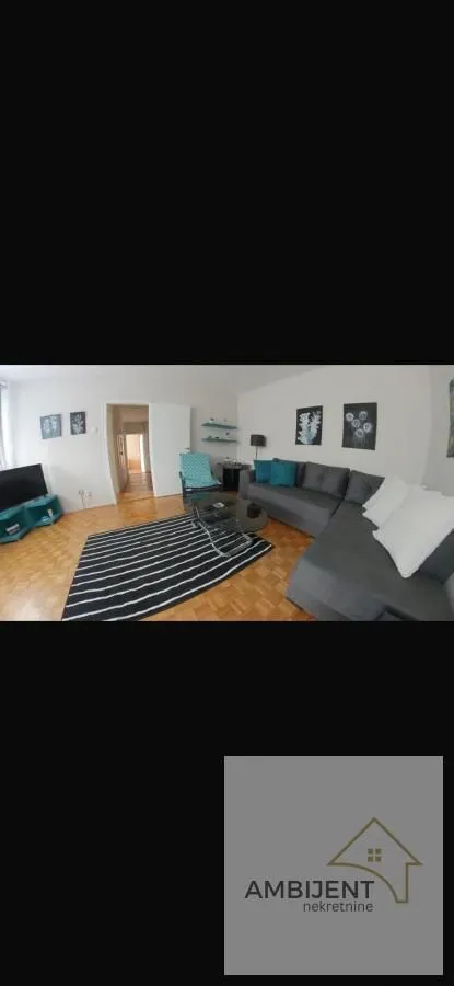 Nameštena dnevna soba sa ugaonom garniturom, televizorom i parketom na podu.