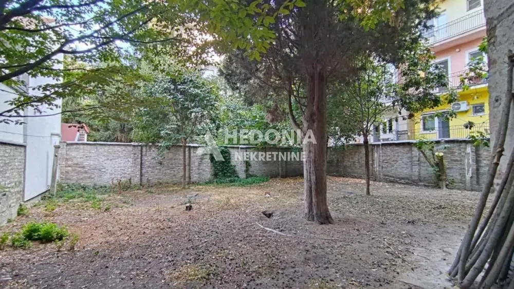 Zagrađeno dvorište sa drvećem i pogledom na okolne zgrade.