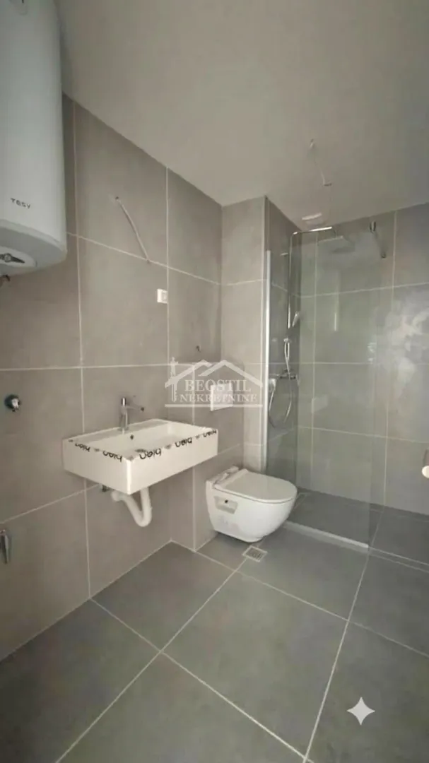 Kupatilo obloženo sivim pločicama sa tuš kabinom, umivaonikom i wc šoljom.