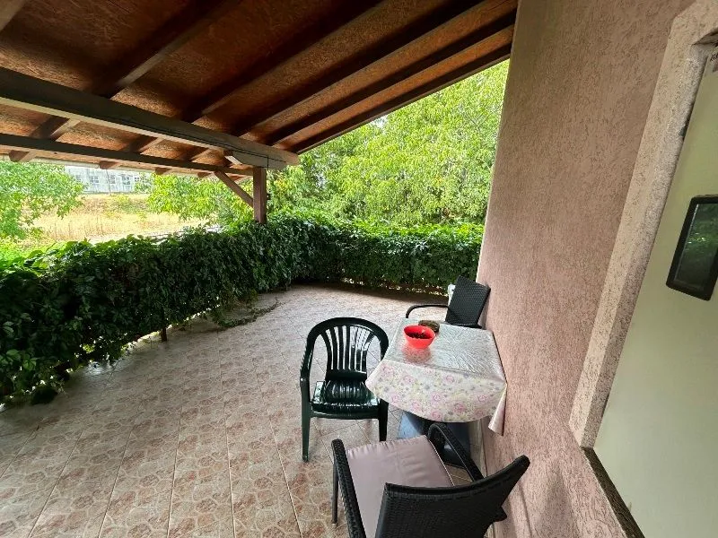 Covered terrace with a table, chairs, and a view of greenery.