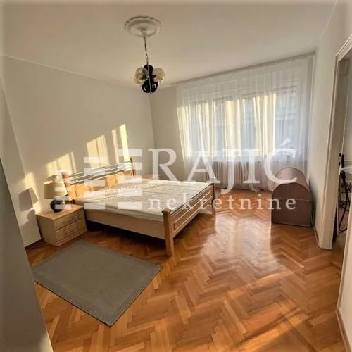 Furnished bedroom with a double bed, wooden parquet flooring, and a large window.