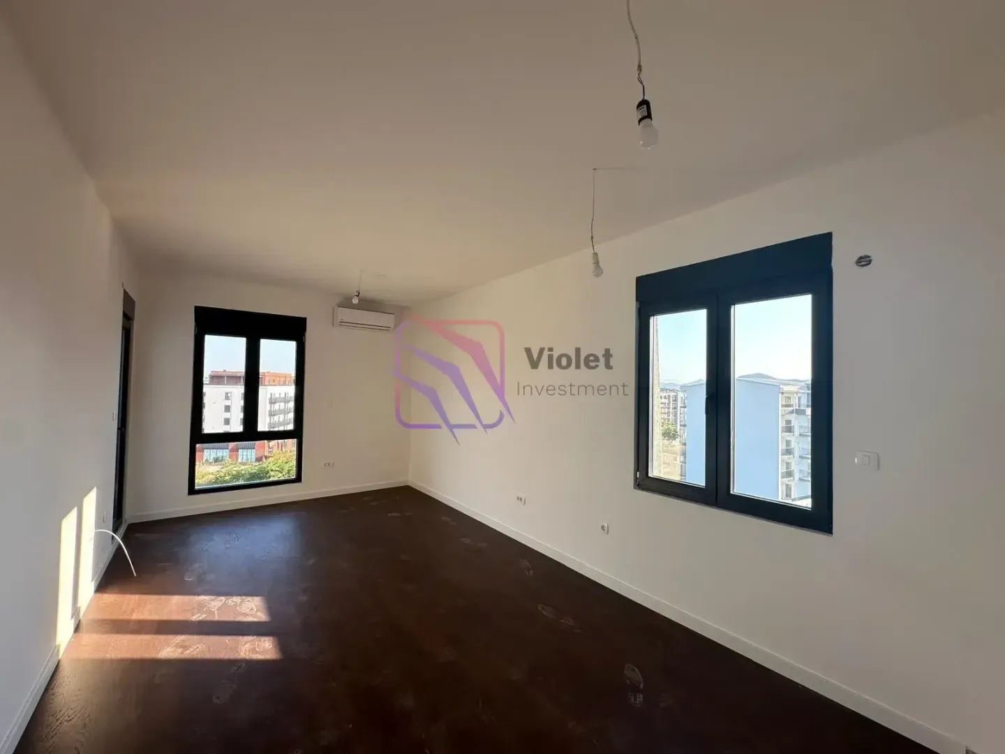 Empty room with white walls, dark hardwood flooring, and windows.