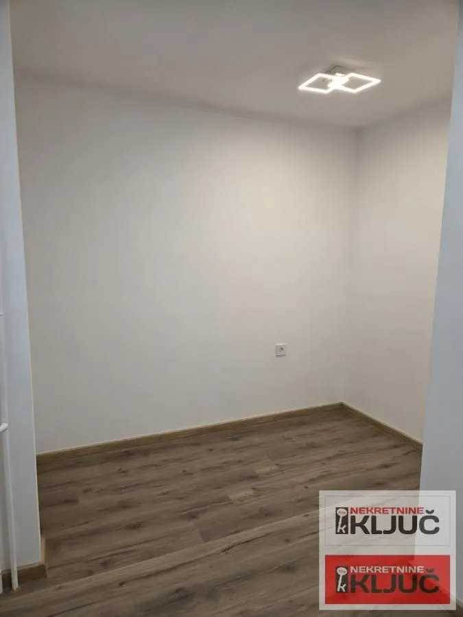 Prazna soba sa belim zidovima, drvenim podom i plafonskom rasvetom.