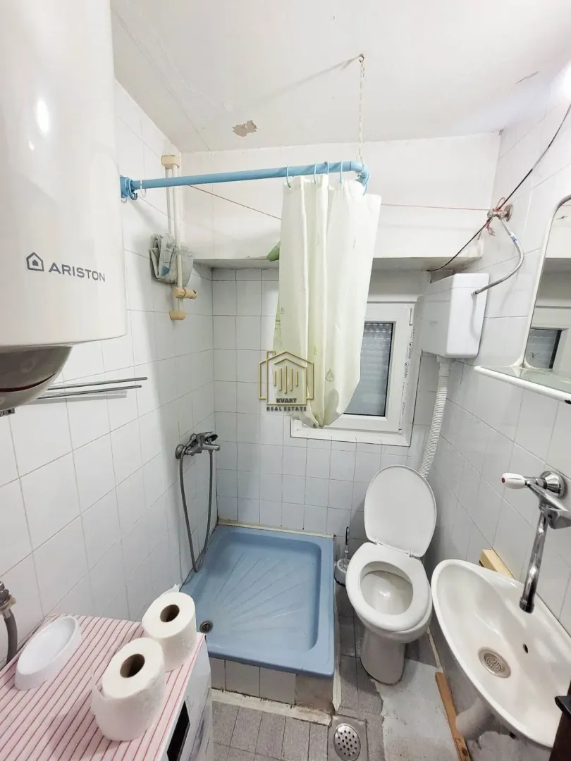 Manje kupatilo obloženo bijelim pločicama sa tuš kadom, wc šoljom i umivaonikom.