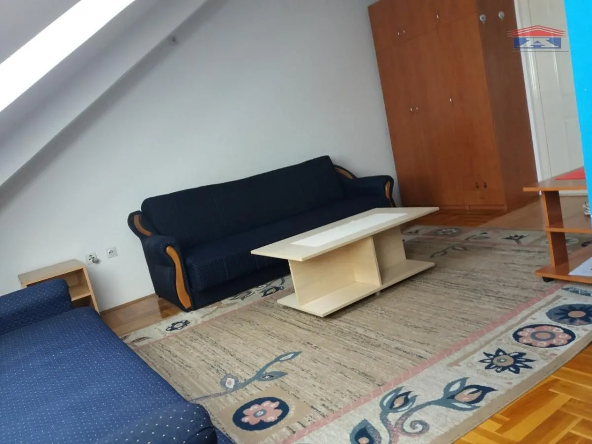 Dnevna soba sa plavim garniturama, stočićem i drvenim ormarom pod kosim krovom.