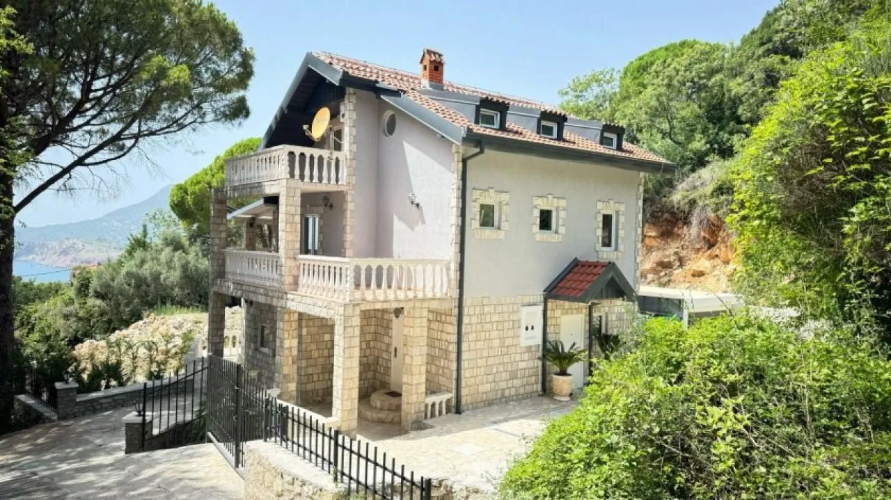 Pogled na spoljašnjost trospratne kuće sa kamenim detaljima i terasama okružene zelenilom.