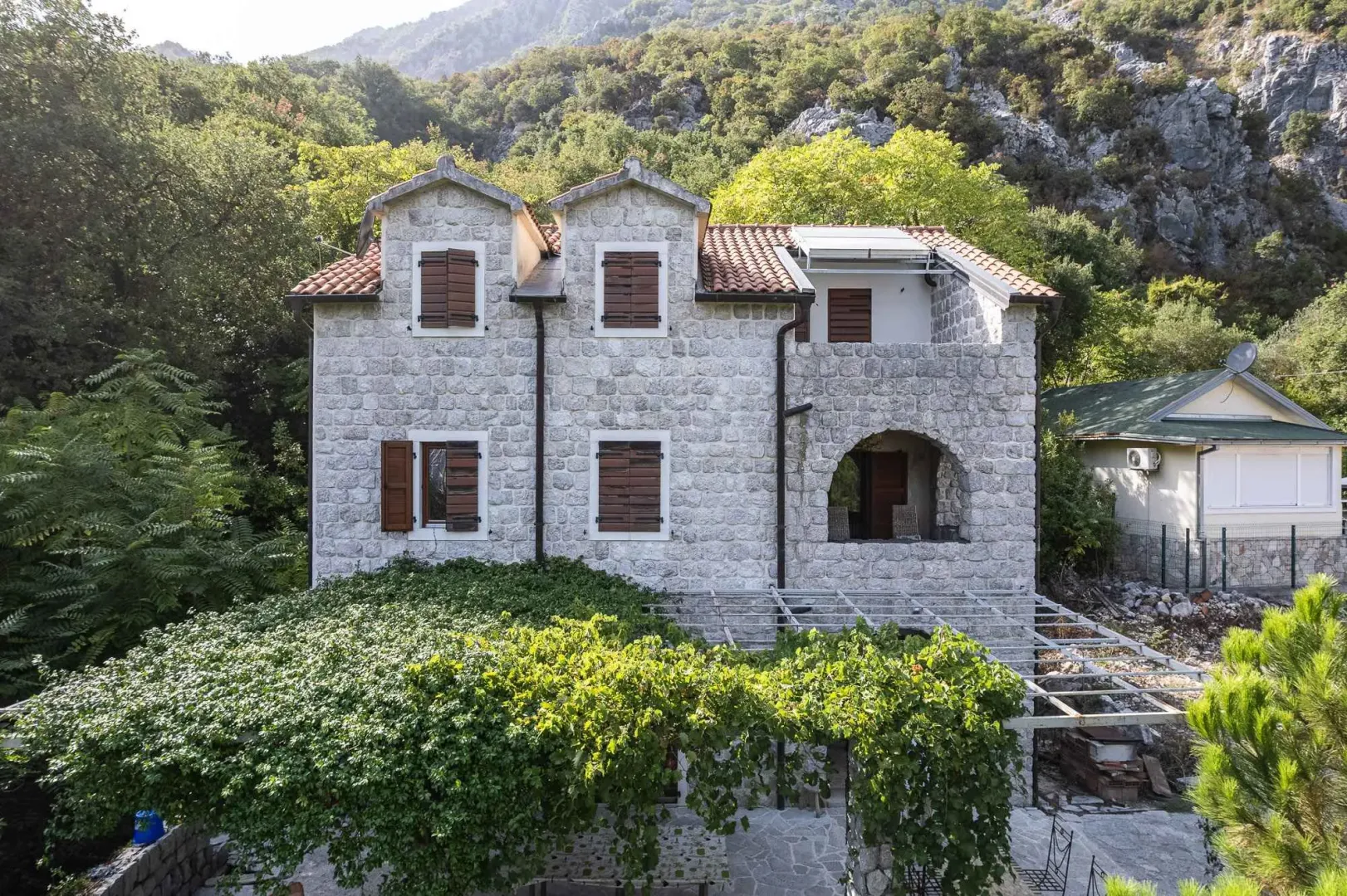 Pogled na prednju fasadu tradicionalne kamene kuće sa drvenim kapcima i zelenilom u dvorištu.