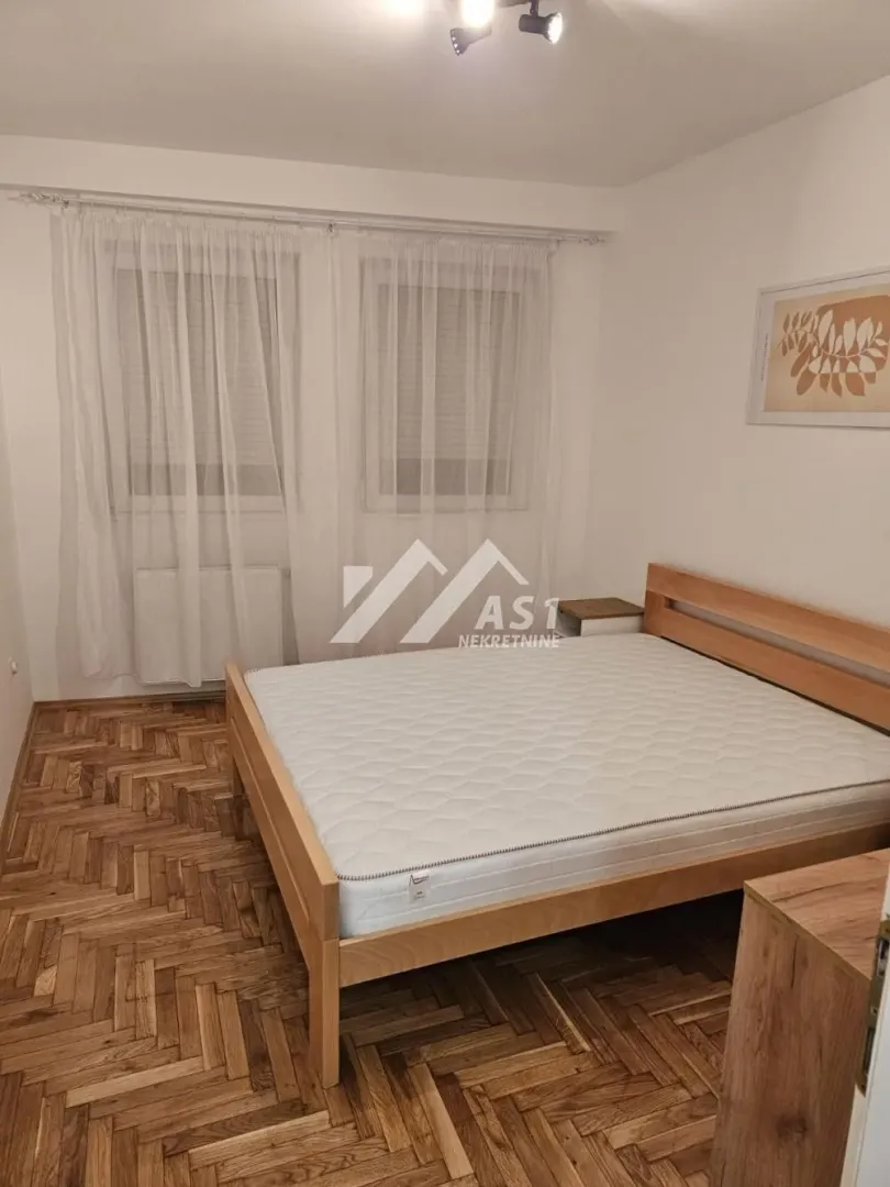 Nameštena spavaća soba sa bračnim krevetom i prozorima sa belim zavesama.