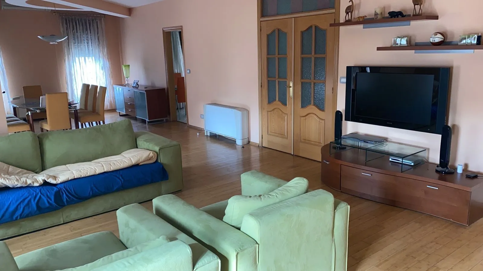 Prostrani dnevni boravak sa zelenim garniturama, televizorom i trpezarijskim stolom u pozadini.
