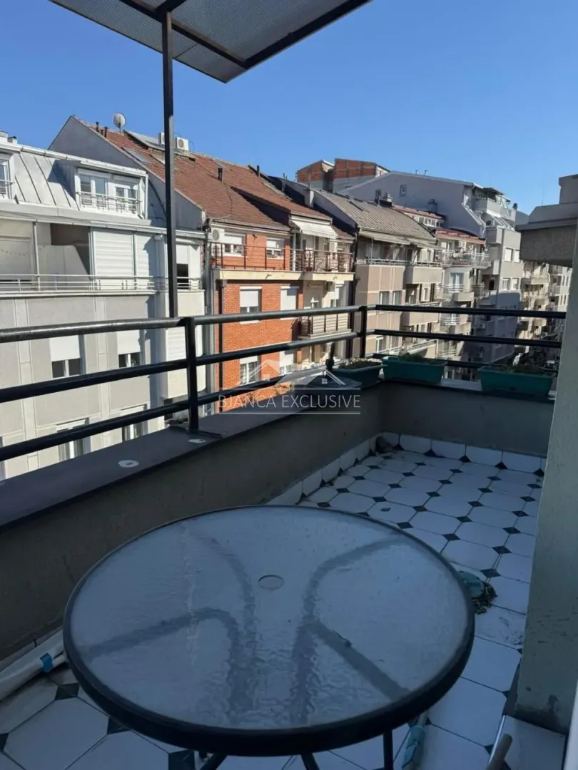 Natkrivena terasa sa stolom i pogledom na okolne zgrade.