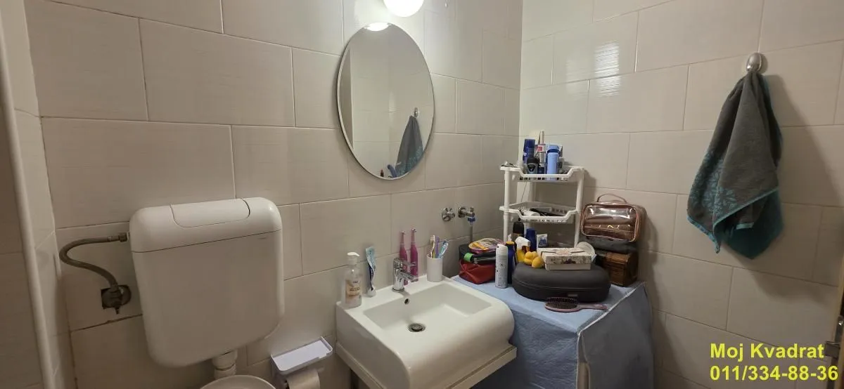 Manje kupatilo sa umivaonikom, ogledalom i WC šoljom obloženo svetlim pločicama.