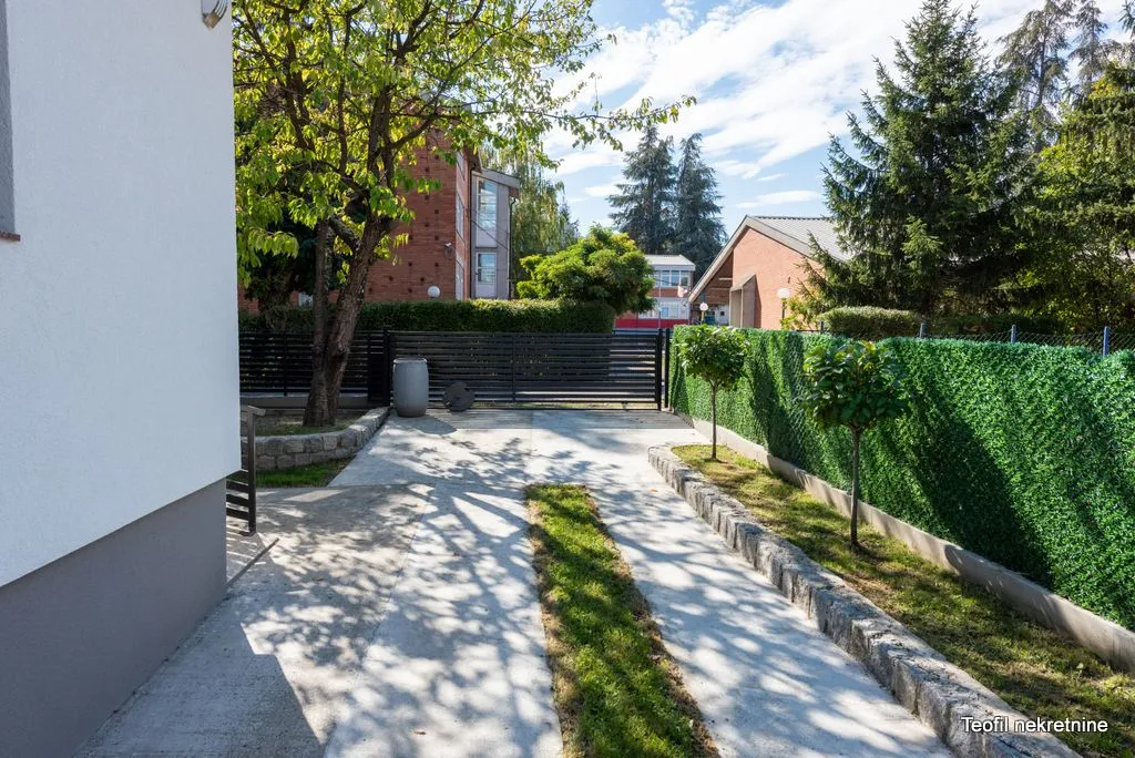 Popločano dvorište sa stazom, zelenilom i ogradom ispred kuća.