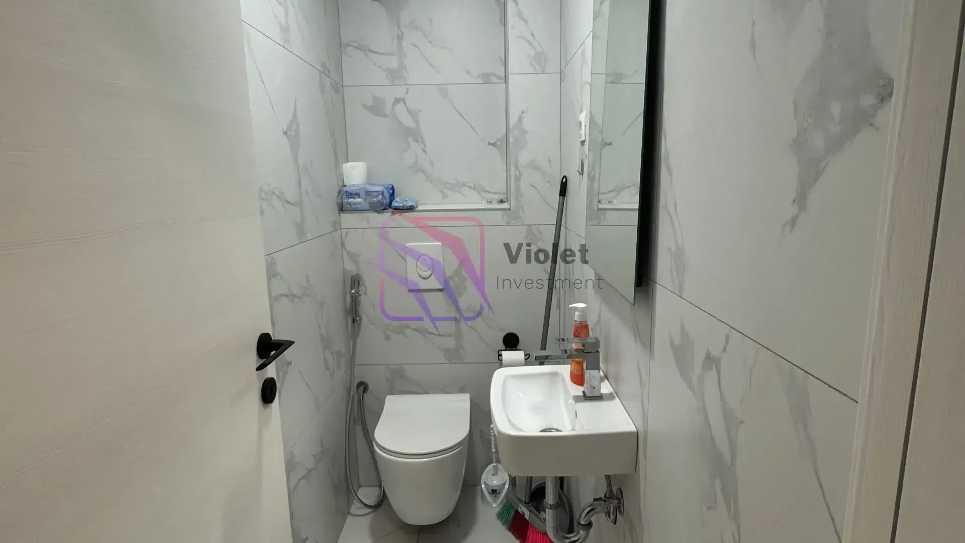 Small bathroom tiled with marble-patterned ceramic tiles, featuring a toilet and a washbasin.