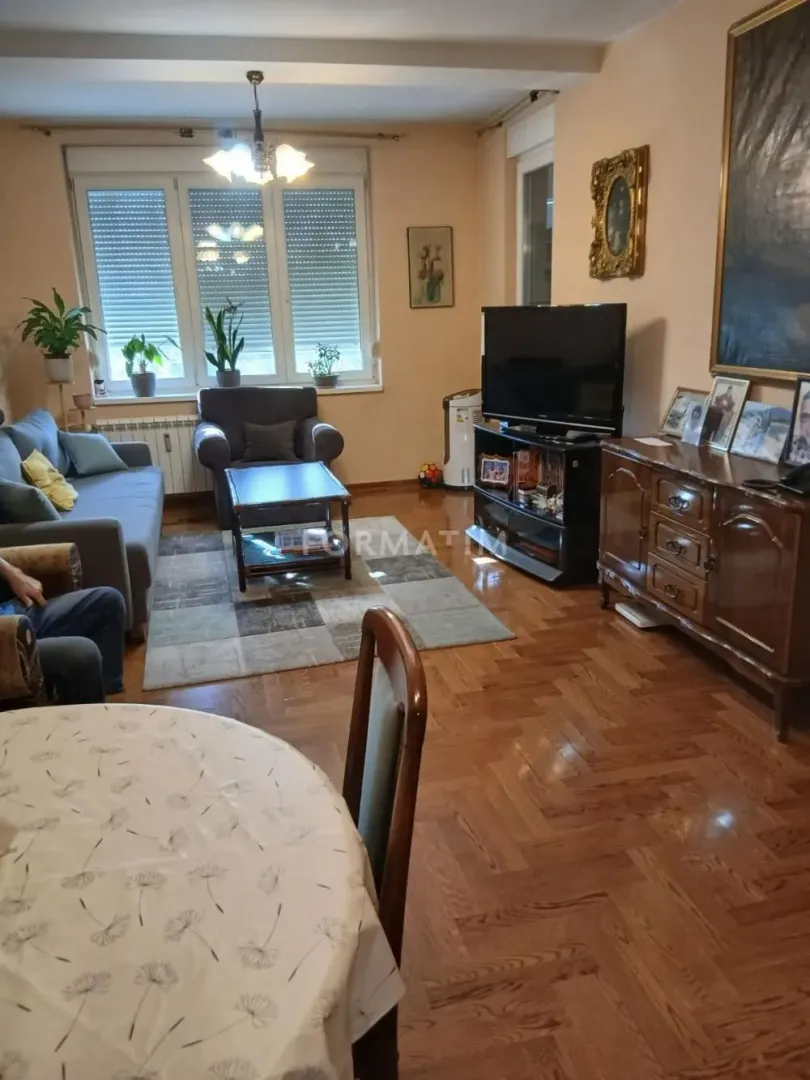 Nameštena dnevna soba sa garniturom, televizorom i trpezarijskim stolom.