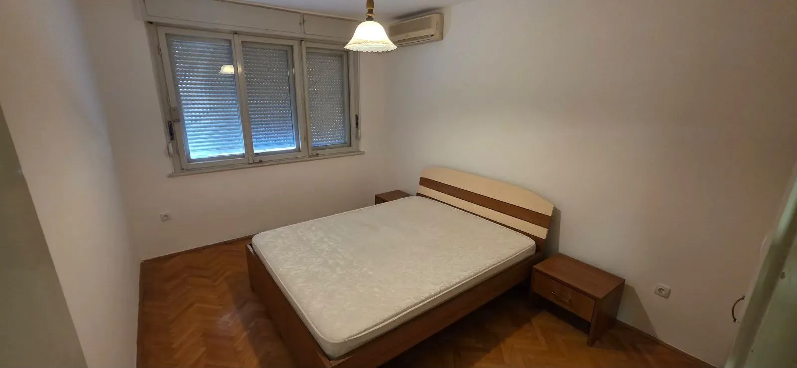 Spavaća soba sa bračnim krevetom, noćnim ormarićima i prozorom sa roletnama.