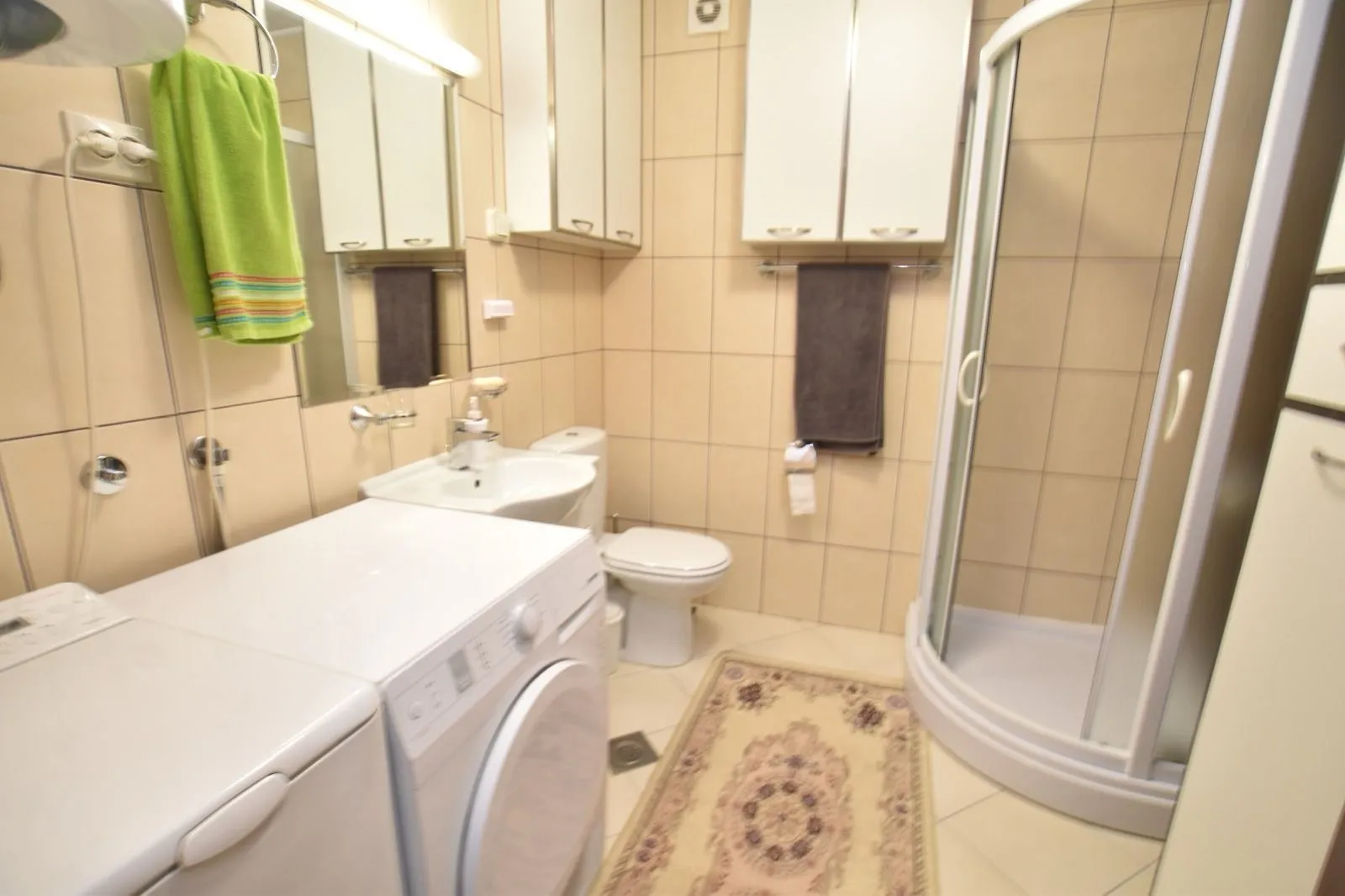 Svijetlo kupatilo sa tuš kabinom, veš mašinom i bež pločicama.