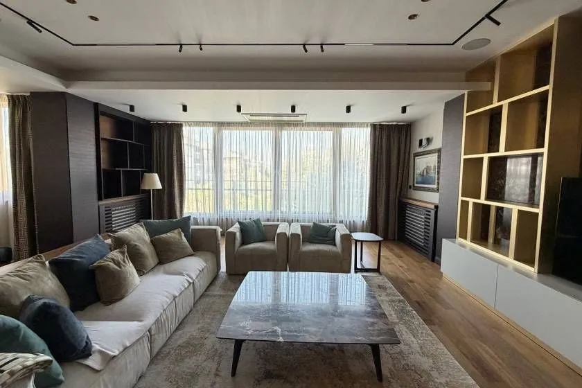 Spacious living room with a sofa set, large windows, and a modern shelving system.