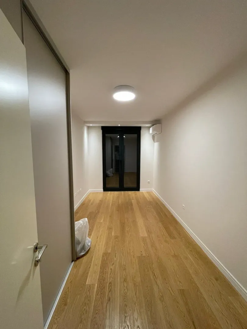 Prazna, osvijetljena soba sa drvenim podom i balkonskim vratima.