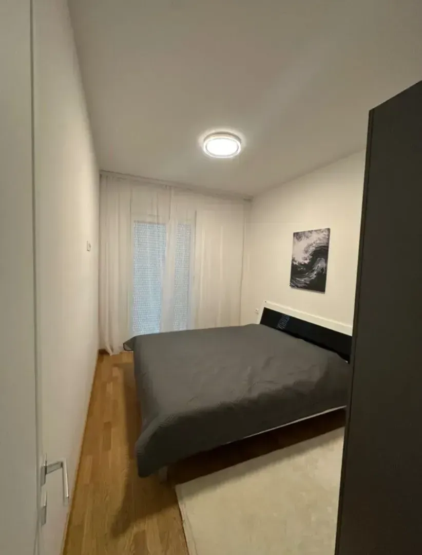 Pogled na namještenu spavaću sobu sa bračnim krevetom i prozorom sa bijelim zavjesama.