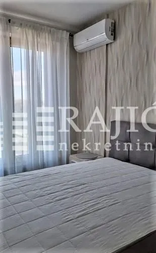 Furnished bedroom with a double bed, air conditioner, and a window with white curtains.
