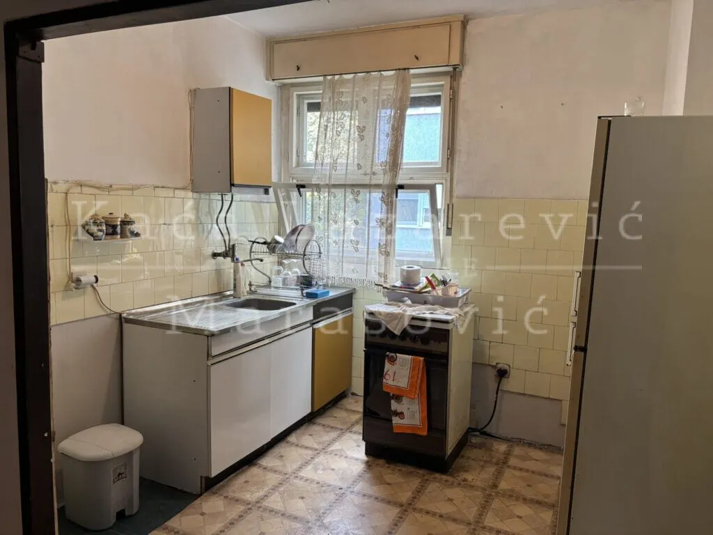 Starija kuhinja sa sudoperom, šporetom i frižiderom pored prozora.