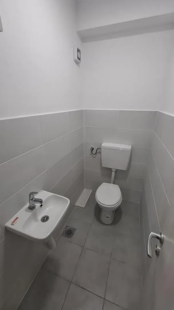 Manje kupatilo sa WC šoljom i umivaonikom obloženo svetlim pločicama.
